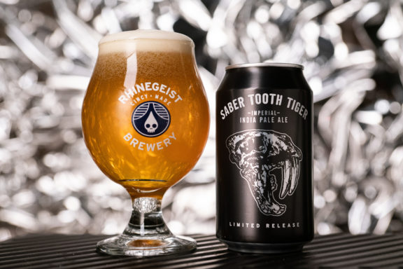 Rhinegeist Saber Tooth Tiger can next to full snifter showing beer